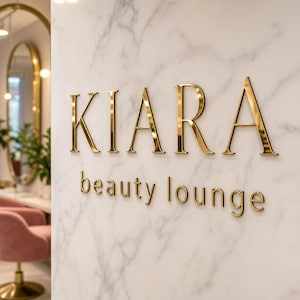 Peut inclure: Un mur en marbre blanc portant le texte métallique doré KIARA beauty lounge dans une police serif. Les lettres sont montées directement sur la surface, créant un effet dimensionnel avec des lignes épurées et une finition polie.