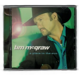 May include: A CD case featuring Tim McGraw. The cover art shows McGraw in a black hat and jacket, with a teal shirt. The album title, "a place in the sun," is displayed below his name in white text.