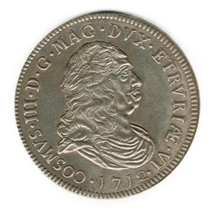 Puede incluir: Una moneda de plata con el perfil de un hombre con pelo largo y bigote. La moneda tiene texto alrededor del borde, incluida la fecha 1712. La moneda es redonda con un borde texturizado y sobre un fondo blanco.