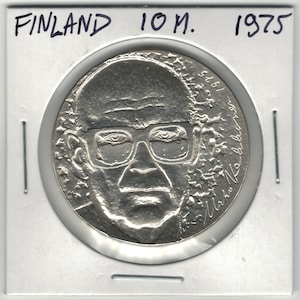 May include: A silver coin from Finland, dated 1975, featuring a portrait of a man wearing glasses. The coin is encased in a clear plastic holder, with the word "FINLAND" and the denomination "10 M." visible.