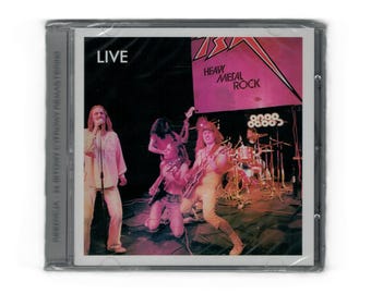 Puede incluir: Una caja de CD con una imagen de una actuación en directo de una banda de heavy metal rock. La portada del álbum tiene la palabra "LIVE" en blanco, con el nombre de la banda y "HEAVY METAL ROCK" en rosa. Los miembros de la banda están en el escenario.