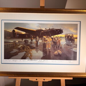 Puede incluir: Una impresión enmarcada que representa un avión antiguo en un aeródromo mojado, con figuras vestidas de época. La obra de arte está enmarcada en un marco dorado, montada sobre un caballete. La escena evoca una sensación de historia y aviación.