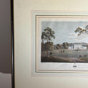May include: Framed landscape print featuring a stately white manor house, trees, and grazing animals. The artwork is set within a cream-colored mat and a black and gold frame. The print includes text at the bottom, indicating the location and title.