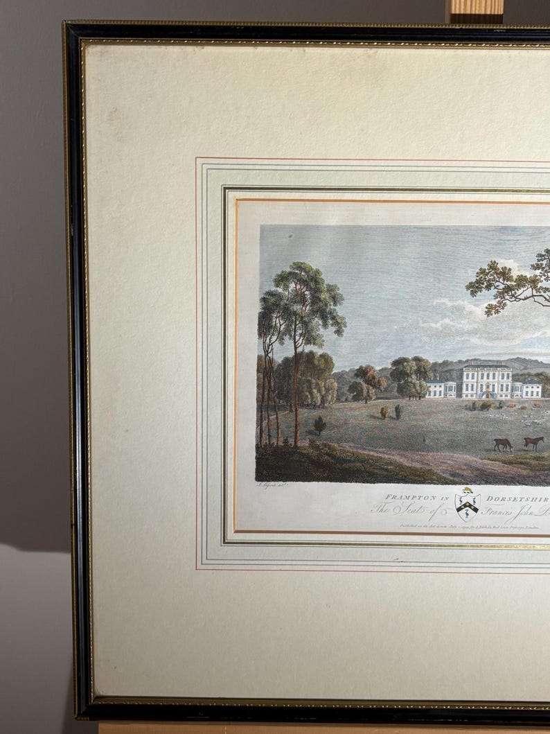 May include: Framed landscape print featuring a stately white manor house, trees, and grazing animals. The artwork is set within a cream-colored mat and a black and gold frame. The print includes text at the bottom, indicating the location and title.
