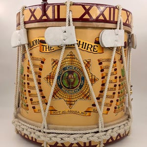 Op de afbeelding: Een vintage trommel met een crèmekleurige body, versierd met de tekst "The Cheshire Regiment" en diverse gevechtslocaties. De trommel heeft een touwspansysteem, leren riemen en een decoratieve bordeauxrode rand.