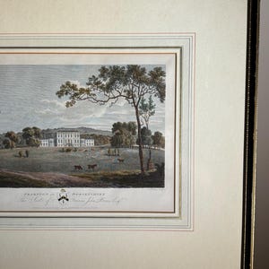 May include: Framed landscape print featuring a stately manor house in a pastoral setting. The artwork, with a cream-colored mat and black frame, depicts a field with trees, deer, and a distant building. The print includes text at the bottom, indicating the location.