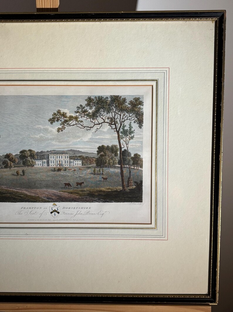 May include: Framed landscape print featuring a stately manor house in a pastoral setting. The artwork, with a cream-colored mat and black frame, depicts a field with trees, deer, and a distant building. The print includes text at the bottom, indicating the location.