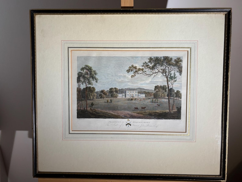May include: Framed antique print of a stately home in a landscape. The artwork features a large white building with multiple windows, surrounded by trees and a grassy field. The print is in a black and gold frame.