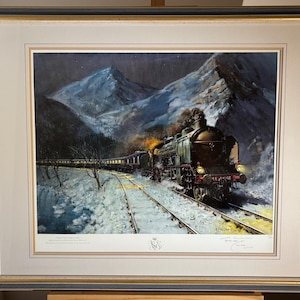 May include: Framed print of a vintage train traveling through a snowy mountain landscape at night. The train is dark green with yellow headlights and smoke billowing from the engine. The artwork is framed in a gold and black frame.
