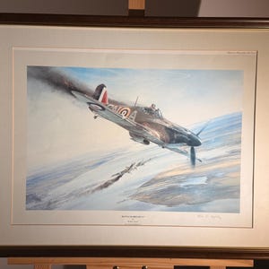 May include: Framed print of a vintage airplane in flight, with a brown wooden frame. The artwork depicts a detailed illustration of a fighter plane with a red and white tail, soaring through a cloudy sky. The print is mounted on a white mat.