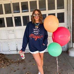 May include: A navy blue sweatshirt with the text "The World Needs Your Magic" in red and white. The person is wearing white shorts with a lace trim, brown boots, and sunglasses, holding a drink and balloons.