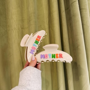 May include: Two off-white hair claw clips, each with the word "MOTHER" in colorful letters. One clip is open, the other is closed. The background is a light green curtain.
