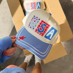 May include: A cream-colored baseball cap with a light blue brim featuring two white stripes. The front panel displays the letters U, S, and A as individual patches with red, white, and blue patterns. A small red star is embroidered on the brim.