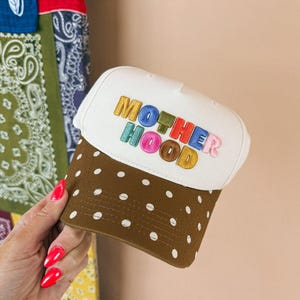 May include: A white baseball cap with a brown brim featuring white polka dots. The front of the cap displays the word MOTHERHOOD embroidered in multi-colored, puffy, block-style letters including yellow, blue, green, red, and pink tones.