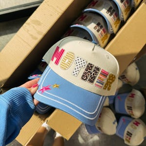 May include: A light blue and cream-colored baseball cap with the word "MOTHER" in patchwork letters. The brim is light blue with white stitching and star accents. Several caps are visible in a cardboard box.
