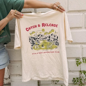 May include: A cream-colored short-sleeve t-shirt featuring a graphic print of a person fishing in a lake with mountains in the background. The text "CATCH & RELEASE" appears above the illustration, and "ETTA & EAST ADVENTURE CLUB" is printed below.