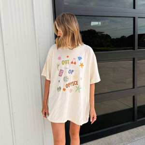 May include: An off-white short-sleeve t-shirt featuring the text "OUT OF OFFICE" in colorful letters. The design includes small graphic illustrations of a palm tree, beach towel, life ring, flip-flops, bikini top, starfish, and various flowers.