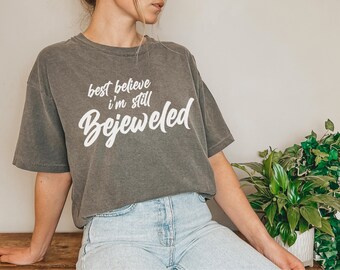 May include: A gray t-shirt with the text "best believe I'm still Bejeweled" in white script. The shirt has a relaxed fit and a vintage wash appearance. The model is wearing light blue jeans.