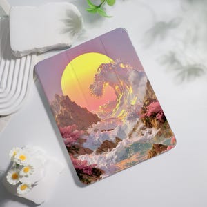 Puede incluir: Una funda rectangular para tableta con un estampado gráfico vibrante de una gran ola oceánica rompiendo contra acantilados rocosos bajo un sol amarillo brillante. La escena incluye árboles de flores rosas a lo largo de la costa.