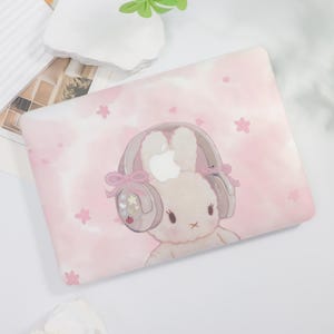 May include: Pink laptop case featuring a cartoon bunny wearing headphones with an Apple logo. The bunny is set against a pink watercolor background with scattered pink flower accents. The case has a smooth, glossy finish.