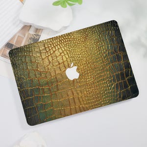 May include: A laptop skin with a textured, gold and dark green crocodile pattern. The skin features a white Apple logo in the center. The design has a gradient effect, transitioning from gold to dark green.