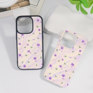 May include: Two phone cases with a floral design. The cases feature a light cream background with purple flowers and green leaves. One case has a black border, while the other is frameless. The design is repeated on both cases.