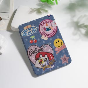 May include: A denim-textured case adorned with embroidered patches. Patches include a smiley face, cherries, a star, and text that reads "Call me," "Craft Hoarder," and "Sailor Moon." The case has a rounded rectangular shape.