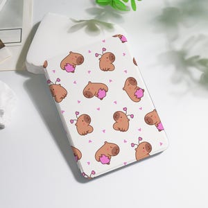May include: A white tablet case featuring a repeating pattern of cartoon capybaras holding pink hearts, with small pink heart accents. The capybaras are brown with small antennae. The case is rectangular and appears to be made of a smooth material.