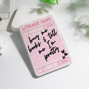 May include: A pink library card design with the text "buy me books & tell me I'm pretty" in black script. The card has a vintage aesthetic with a grid pattern and heart accents. The card also has the text "LIBRARY CARD" and the number "026483".