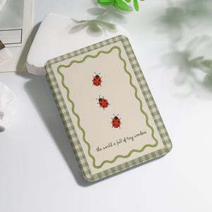 May include: A rectangular, cream-colored coaster with three red ladybugs and the text "the world is full of tiny wonders." The coaster has a green and white checkered border and a scalloped edge.