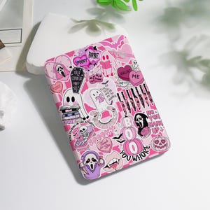 May include: Pink and white Halloween-themed phone case covered in stickers. The stickers include ghosts, pumpkins, hearts, and text like "Spooky", "Haunt Me", and "You Whore". The case has a checkered pattern and a variety of spooky graphics.