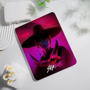 May include: A tablet case featuring a digital illustration of a person with blue skin, glowing yellow eyes, and a wide-brimmed hat. The background is a gradient of pink and purple. The text "SAJA BOYS" is in white and pink.
