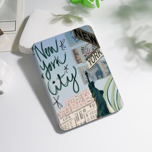 Könnte beinhalten: Eine rechteckige Handyhülle mit New York City-Motiv. Das Design zeigt die Freiheitsstatue, Straßenschilder und die Worte "New York City" in handschriftlichem Stil. Der Hintergrund ist hellblau mit weiteren New York City-Bildern.