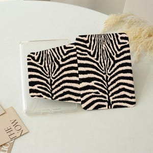 May include: A rectangular protective case for an electronic device featuring a black and beige zebra stripe pattern. The case has a clear plastic frame and a textured, patterned front panel that displays a bold, repeating animal print design.