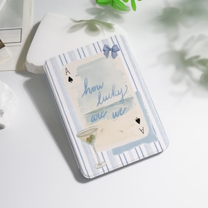 May include: A rectangular case with a blue and white striped pattern. The case features a watercolour illustration of a martini glass, the Ace of Spades, and the text "how lucky are we". A blue bow is at the top.