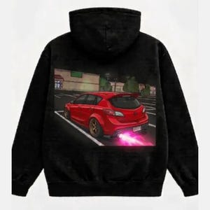 May include: Black hoodie with a graphic of a red hatchback car with a pink exhaust trail, parked in a lot. The car has gold rims and a spoiler. The background shows a building and trees.