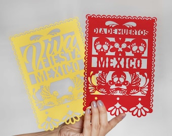 May include: Two paper cutouts, one yellow and one red, with traditional Mexican designs. The yellow cutout says "Viva Fiesta Mexico" and the red cutout says "Dia de Muertos Mexico" with two skull designs.