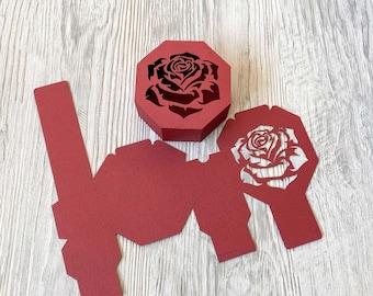 Könnte beinhalten: Eine rote Papierbox mit Rosenmuster. Die Box ist zusammengebaut und bereit, mit einem Geschenk gefüllt zu werden. Die Box ist aus Karton und hat ein filigranes, kompliziertes Design.