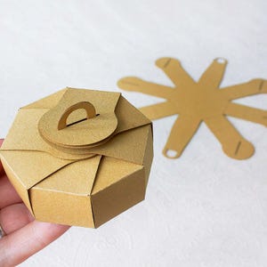 Op de afbeelding: Een bruine kartonnen doos met een handvat aan de bovenkant. De doos is gevouwen in een ronde vorm en heeft een stervormige uitsparing aan de zijkant.