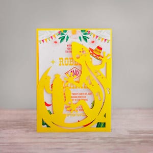 Puede incluir: Invitación amarilla con un diseño recortado de un lagarto con un sombrero. El texto de la invitación dice: "Con ustedes a la boda de Robert y Maria, veintinueve de junio, dos mil diecinueve, fiesta por la noche, 10000 Ula Place, Pakal, FL 33074, recepción a continuación."