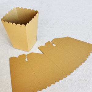 May include: A brown cardboard box with a scalloped edge. The box is flat and unfolded, showing the creases and folds that will form the box when assembled. The box is designed to hold small items, such as gifts or treats.