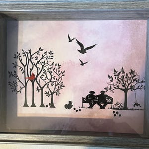 May include: A framed print with a pink and purple watercolor background. The print features a silhouette of two people sitting on a bench, a red cardinal perched on a tree branch, and several trees. There are also silhouettes of birds flying in the sky.
