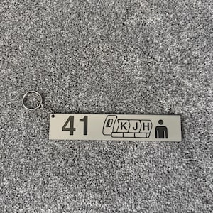 Puede incluir: Un llavero rectangular plateado con el n&uacute;mero "41" y un gr&aacute;fico de asiento de tren. El llavero tambi&eacute;n presenta las letras "D K J H" y una figura de palo. Est&aacute; unido a un anillo plateado.