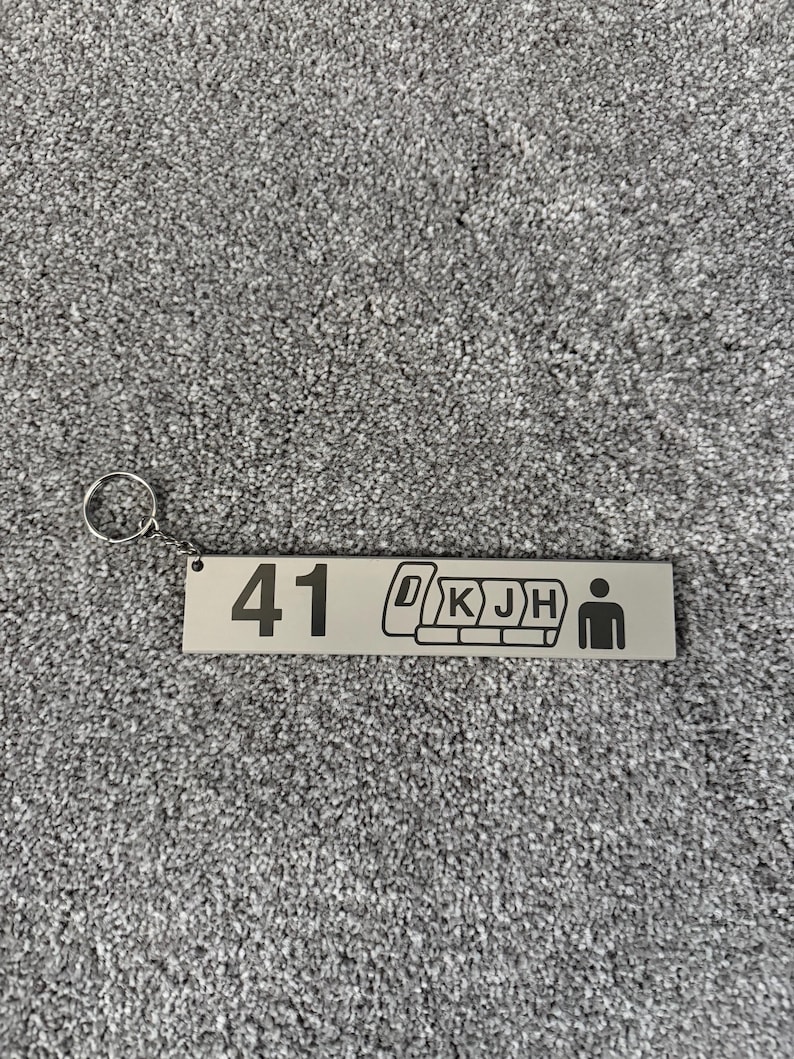 Puede incluir: Un llavero rectangular plateado con el n&uacute;mero "41" y un gr&aacute;fico de asiento de tren. El llavero tambi&eacute;n presenta las letras "D K J H" y una figura de palo. Est&aacute; unido a un anillo plateado.
