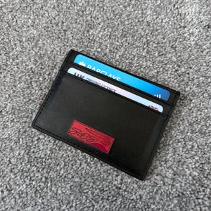 May include: Black leather card holder with a red logo. The card holder contains a Barclays credit card and a UK driving license. The logo is a stylized design with the numbers 747.