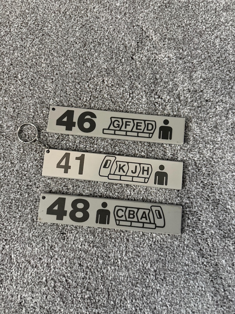 Puede incluir: Tres llaveros rectangulares grises con n&uacute;meros y s&iacute;mbolos negros. Cada llavero tiene un n&uacute;mero, un gr&aacute;fico de disposici&oacute;n de asientos y un icono de persona. Un llavero tiene un anillo de metal adjunto.
