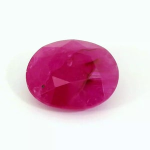 May include: A faceted, oval-shaped ruby gemstone. The gem is a vibrant shade of pinkish-red, with visible internal inclusions and facets that catch the light. The gemstone is set against a plain white background.