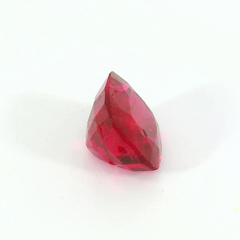 May include: A faceted, teardrop-shaped gemstone in a vibrant shade of red. The gem is set against a plain white background, highlighting its polished surfaces and internal clarity. The cut of the stone creates multiple reflective facets.