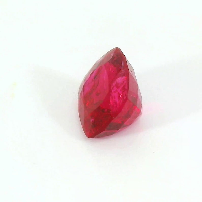 May include: A faceted, marquise-cut ruby gemstone. The gem is a deep, vibrant red color and has multiple facets that reflect light. The gemstone is set against a white background.
