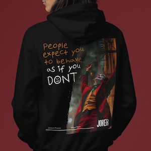 May include: Black hoodie with a graphic print of the Joker from the movie Joker. The text "People expect you to behave as if you don't" is written on the hoodie. The text "Joaquin Phoenix" and "Arthur Fleck" are also printed on the hoodie.